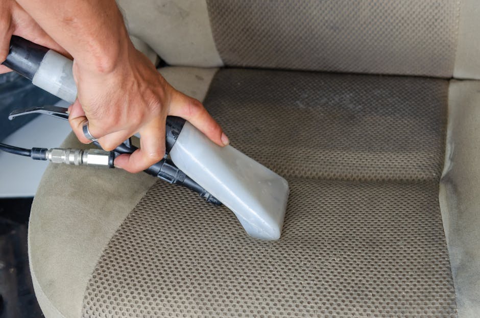 Close-up of a person's hand holding a vacuum cleaner attachment, vacuuming a lime green upholstered armchair with tufted detailing in a well-lit living room. Behind the armchair, there is a dark gray